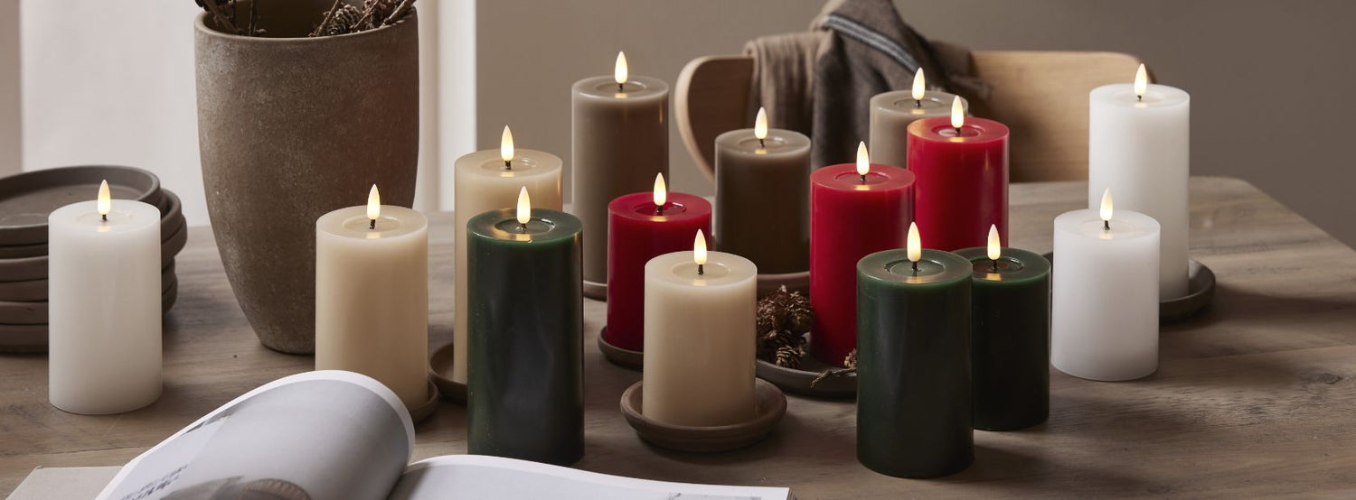 Többszínű LED gyertyák – fehér, bézs, bordó, zöld és szürke árnyalatokban – egy fa asztalon elrendezve, természetes és ünnepi dekorációként.