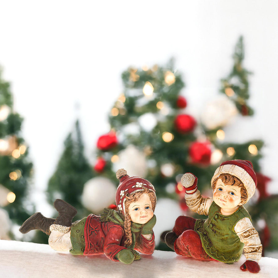 Two Heritage Christmas winter figurines lying on snowy ground with blurred christmas trees in the background