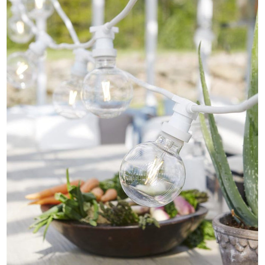 Outdoor garden setting with white round glass bulb string lights above a dining table and potted plants.