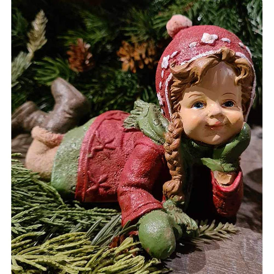 Vintage stílusú, 19,5 cm hosszú, tradicionális megjelenésű, karácsonyi téli sapkás kislány sállal a nyakában zöld fenyőfa előtt