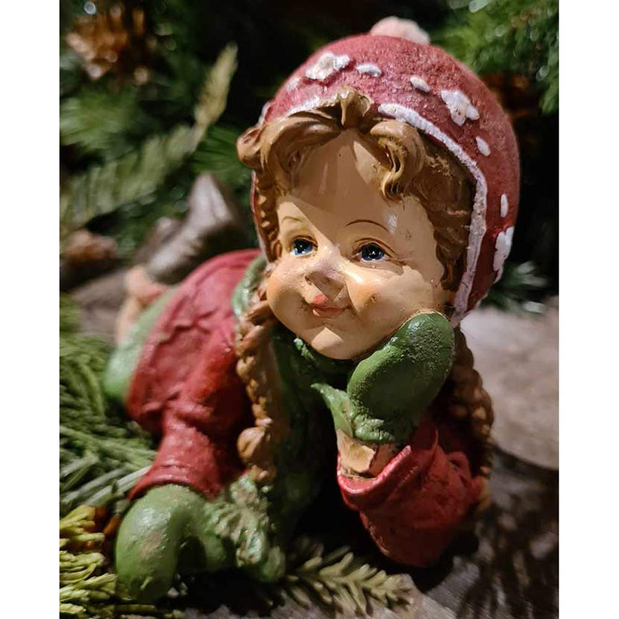 Vintage stílusú, 19,5 cm hosszú, tradicionális megjelenésű, karácsonyi téli sapkás kislány sállal a nyakában zöld fenyőfa előtt