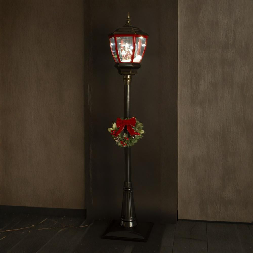 Megvilágított karácsonyi állólámpa sötét háttér előtt – látható a téli falu jelenet és a koszorú.