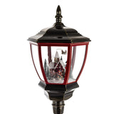 Piros keretes lámpásfej részlet – belül havas ház, fenyőfák és Mikulás szánja a téli jelenetben.