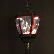 Megvilágított téli falu jelenet közelről – havas ház előtt figurák, fenyőfák és utcai lámpa.