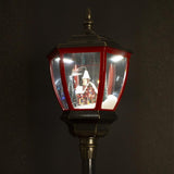 Megvilágított téli falu jelenet közelről – havas ház előtt figurák, fenyőfák és utcai lámpa.