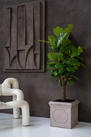 Heritage matt taupe klasszikus fiberstone kaspó lantlevelű fikusszal, kortárs nappaliban, fehér buklé fotel és geometrikus faldekoráció mellett.