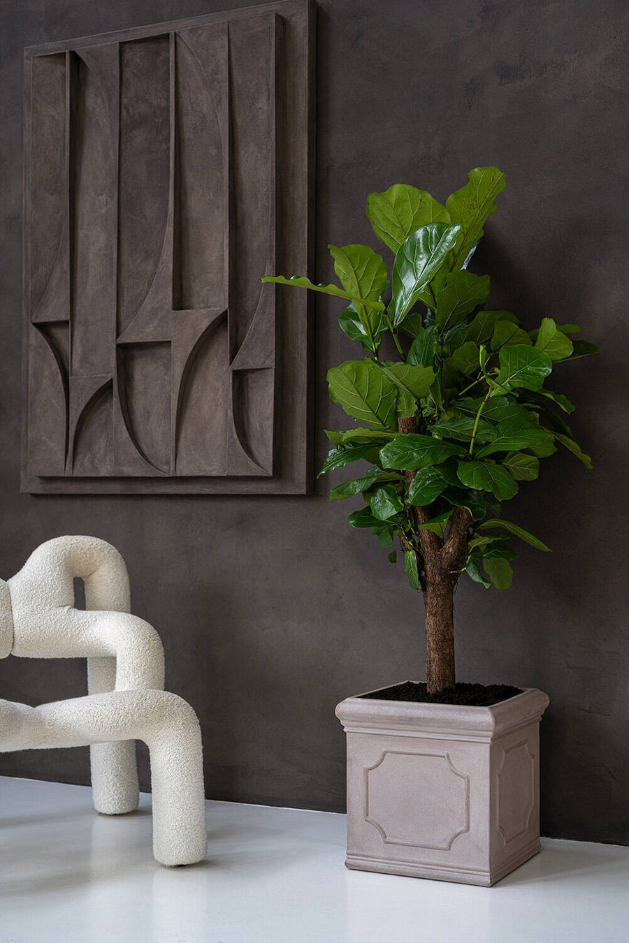 Heritage matt taupe klasszikus fiberstone kaspó lantlevelű fikusszal, kortárs nappaliban, fehér buklé fotel és geometrikus faldekoráció mellett.