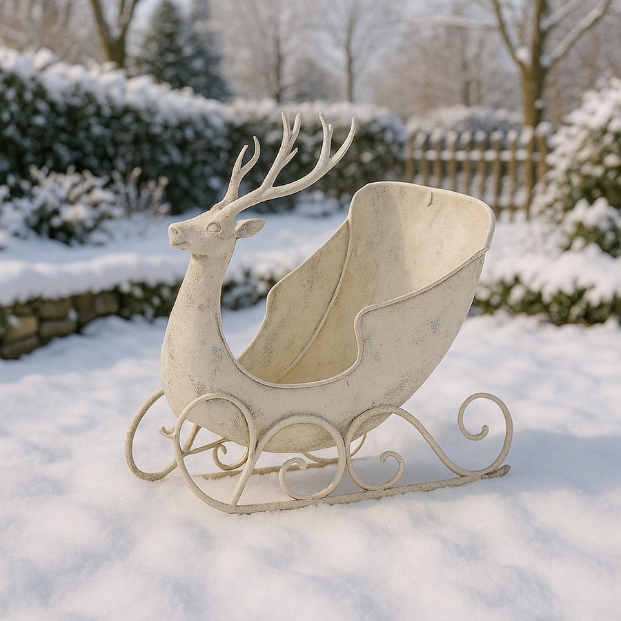 Antikolt krém színű, szarvasfigurás fém dekor szán téli kertben, havas háttérrel, nappali fényben, rusztikus karácsonyi hangulatot idézve.