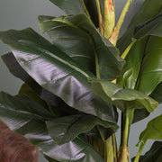 A mű banánfa leveleinek élethű részletei beltéri környezetben, zöld fal és bőrfotel mellett.