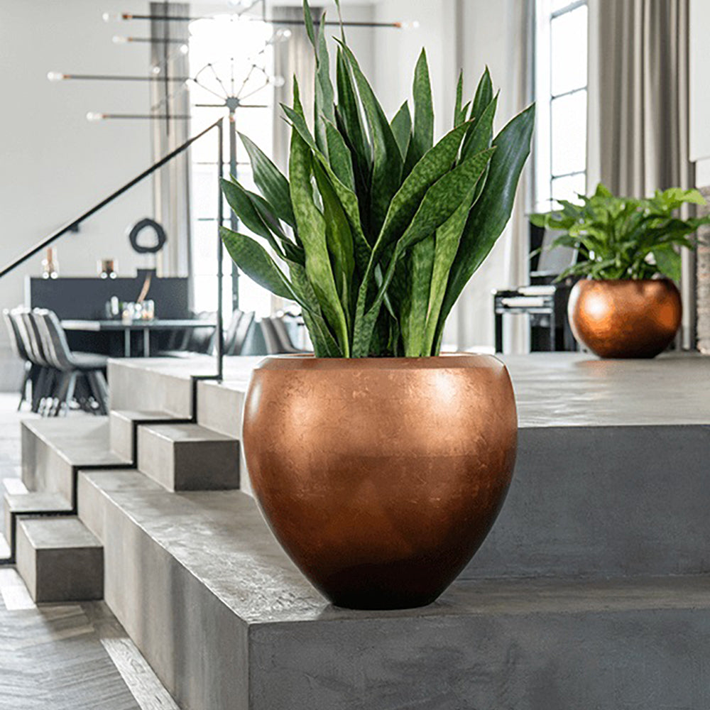 Matt réz színű BAQ METALLIC kompozit óriás kaspó modern enteriőrben, lépcsős betonfelületen elhelyezve, benne dekoratív szobai növénnyel.