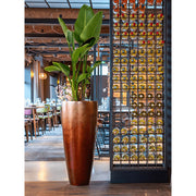 Modern enteriőrben elhelyezett magas, matt réz kaspó buja zöld növénnyel.