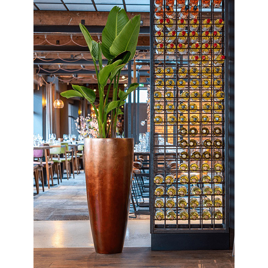 Modern enteriőrben elhelyezett magas, matt réz kaspó buja zöld növénnyel.