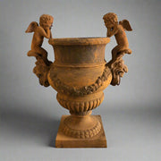 Château Jardin antikolt öntöttvas kaspó puttófigurákkal, műtermi világításban, klasszikus díszítőelemekkel és patinás felülettel.