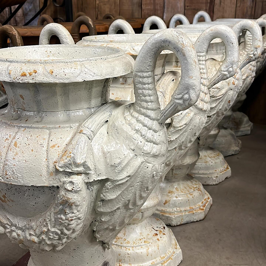 Antikolt öntöttvas kerti díszvázák sorban, hattyúfejes fogantyúval és virágfüzérrel, Château Jardin kollekció, raktári környezetben.