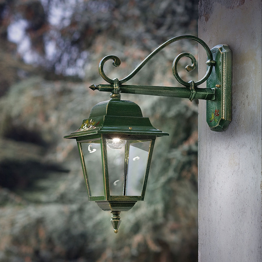 Ferroluce Gorizia klasszikus stílusú falilámpa felszerelve egy rusztikus ház falán, erdei háttérrel, üzem közben világítva.