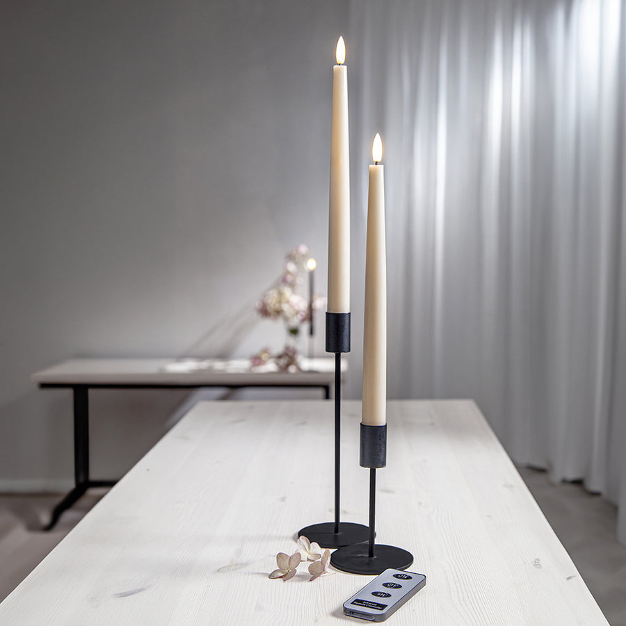 Két bézs LED vacsoragyertya fekete gyertyatartóban, világító lánggal, világos asztalon, mellette távirányító és díszvirág.