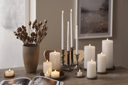 Fehér LED gyertyák különböző méretben és stílusban tálcán és gyertyatartóban, természetes dekorációval berendezett étkezőasztalon.
