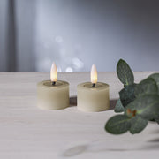 Két bézs színű LED teamécses világít világos faasztalon, elmosódott háttérrel és zöld levél dekorációval.