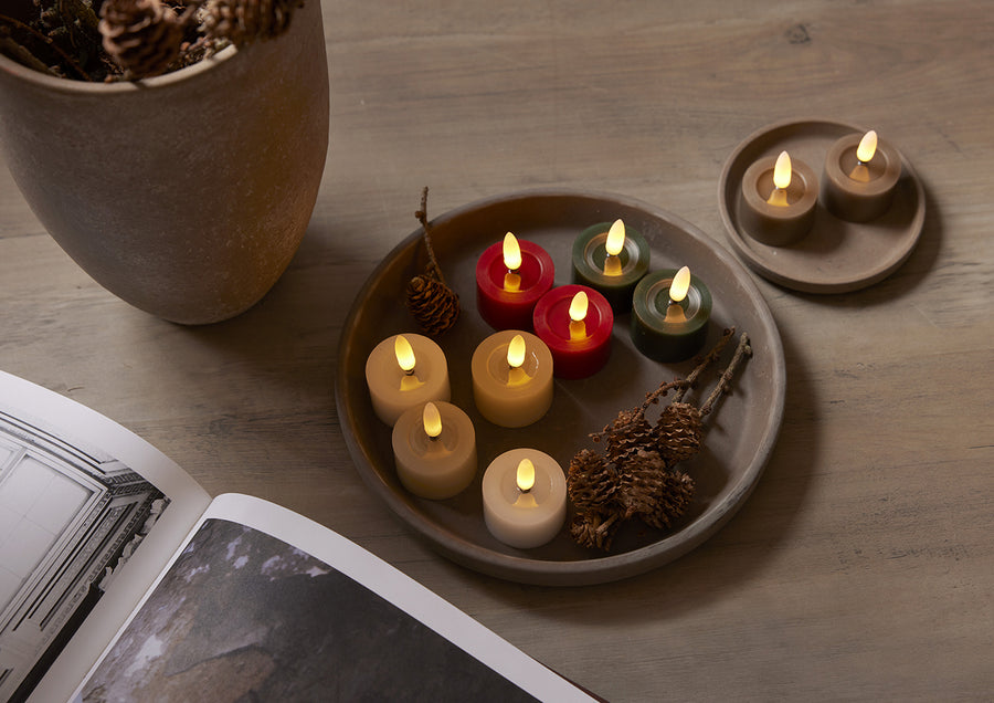 Többszínű Star Trading LED teamécsesek tálcán elrendezve, köztük zöld, piros, bézs és fehér változatok, természetes dekorációval és tobozokkal díszített, hangulatos enteriőrben.