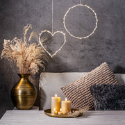 Bézs LED gyertyák és teamécsesek egy arany tálcán elhelyezve, elegáns nappali enteriőrben, szürke fal előtt, díszpárnákkal és fényfüzérrel.