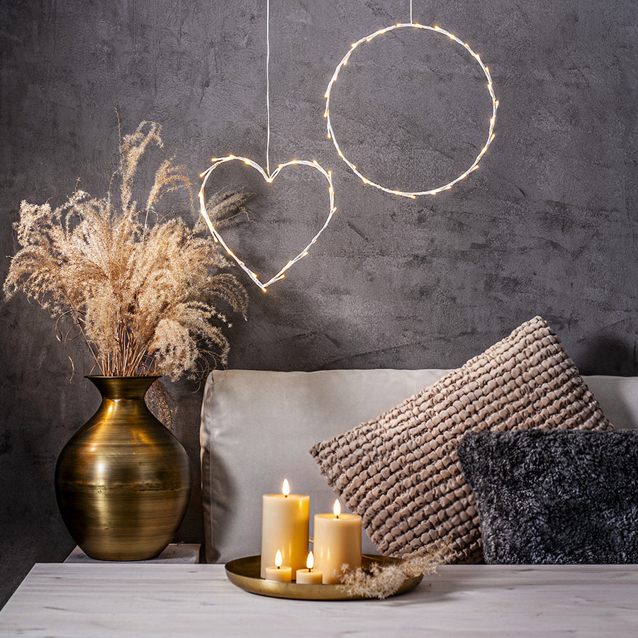 Bézs LED gyertyák és teamécsesek egy arany tálcán elhelyezve, elegáns nappali enteriőrben, szürke fal előtt, díszpárnákkal és fényfüzérrel.