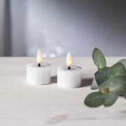 Két fehér színű LED teamécses világít világos faasztalon, elmosódott háttérrel és zöld levél dekorációval.