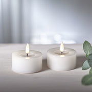 Két fehér színű LED teamécses meleg fehér fénnyel világítva, világos fa felületen, háttérben bokeh fényekkel.