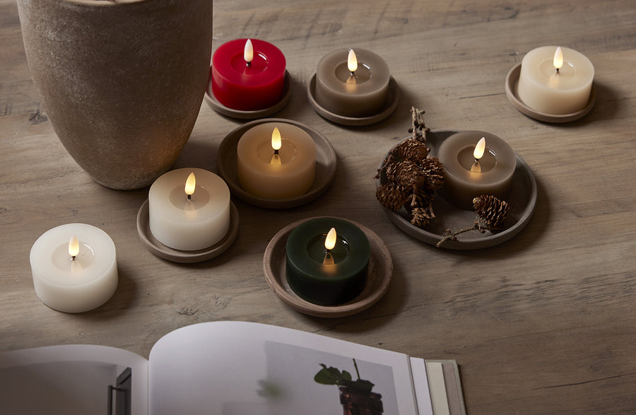 Többféle színű, valódi viaszból készült LED teamécsesek (köztük zöld, fehér, bézs és piros), natúr tálkákon elrendezve egy fa asztalon, fenyőtobozokkal és nyitott magazinnal dekorálva.