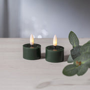 Két zöld LED teamécses meleg fényű lánggal, világos fa felületen elhelyezve, bokeh háttérrel és eukaliptusz ág dekorációval.