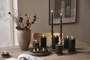 Zöld színű Star Trading LED gyertyák különböző méretekben, természetes hangulatú tálcákon és gyertyatartókban elhelyezve, fa asztalon, skandináv stílusú enteriőrben.