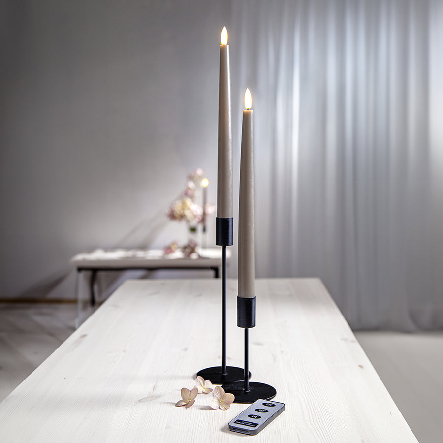 Két Flow szürkésbarna LED vacsoragyertya elegáns fekete gyertyatartóban világos faasztalon, időzítővel és távirányítóval – letisztult, modern enteriőrben.