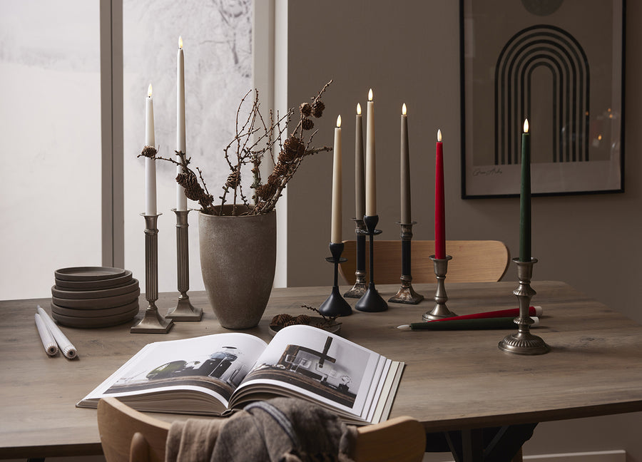Elegáns enteriőr LED vacsoragyertyákkal több színben – fehér, bézs, szürke, bordó és zöld –, klasszikus gyertyatartókban egy természetes hatású faasztalon, háttérben téli ablakkilátással.