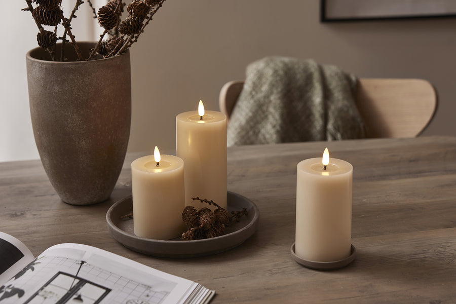 Három bézs LED gyertya valódi viaszból, pislákoló lánggal, természetes dekorációval és meleg enteriőrben, fa tálcán és asztalon elrendezve.