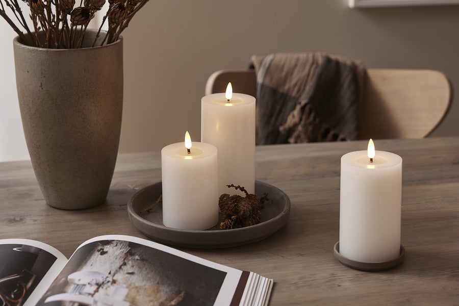 Három különböző méretű, fehér LED gyertya faasztalon tálcára és alátétre helyezve, természetes dekorációval és meleg fehér fényű lánggal.