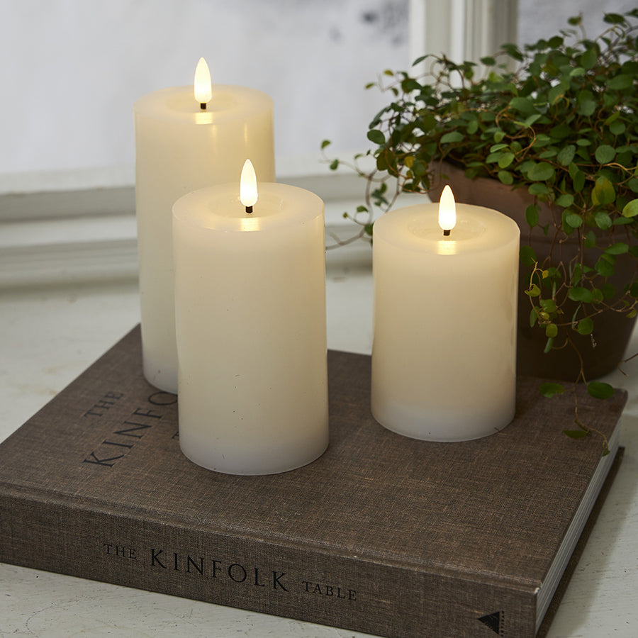 Fehér LED gyertyák a Star Trading Flow kollekcióból, természetes hatású lánggal, dekorációként elhelyezve egy könyvre, növénnyel és ablakpárkánnyal a háttérben,