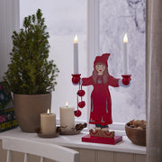 Két fehér LED gyertya egy ablakpárkányon, mellettük egy piros ruhás karácsonyi dekorfigura és egy kis fenyőfa, téli háttérrel.