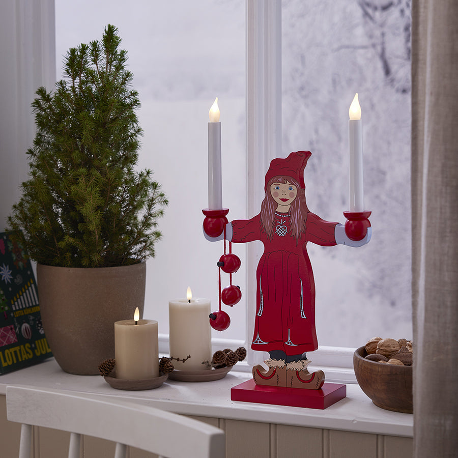 Két fehér LED gyertya egy ablakpárkányon, mellettük egy piros ruhás karácsonyi dekorfigura és egy kis fenyőfa, téli háttérrel.