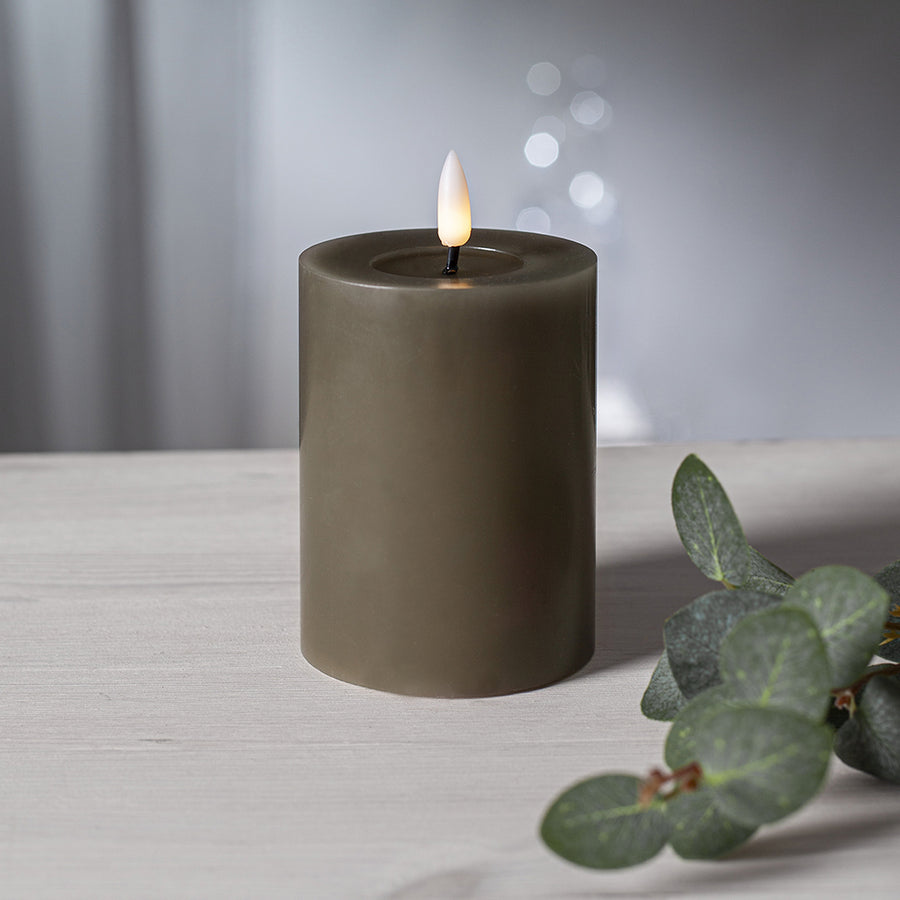 Szürkésbarna LED gyertya meleg fényű lánggal, világos fa asztalon elhelyezve, eukaliptuszág mellett, elmosódott fényfoltokkal a háttérben.