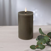 Szürkésbarna LED gyertya meleg fényű lánggal, világos fa asztalon elhelyezve, eukaliptuszág mellett, elmosódott fényfoltokkal a háttérben.