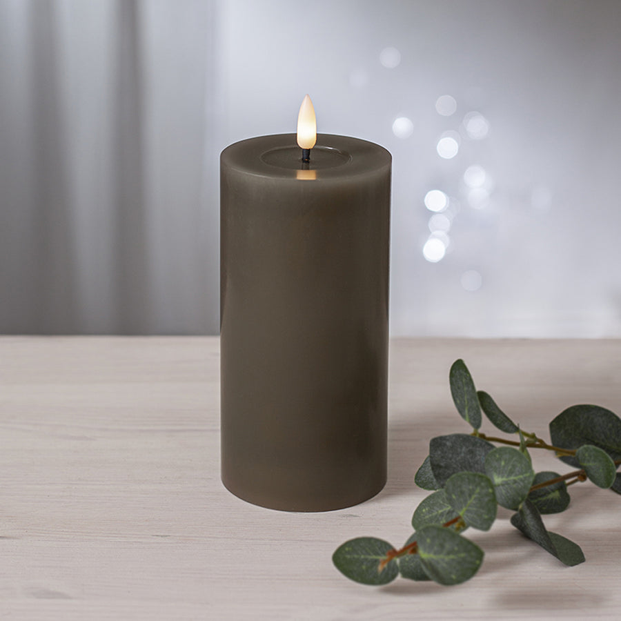 Szürkésbarna LED gyertya meleg fényű lánggal, világos fa asztalon elhelyezve, eukaliptuszág mellett, elmosódott fényfoltokkal a háttérben.