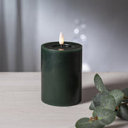 Zöld LED oszlopgyertya valódi viaszból, pislákoló lánggal, világos fa felületen, eukaliptuszlevéllel díszítve.