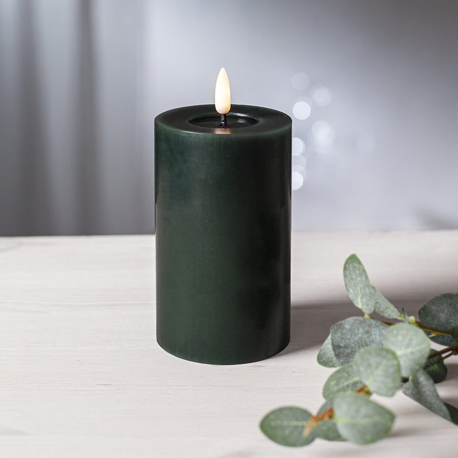 Zöld LED oszlopgyertya valódi viaszból, pislákoló lánggal, világos fa felületen, eukaliptuszlevéllel díszítve.