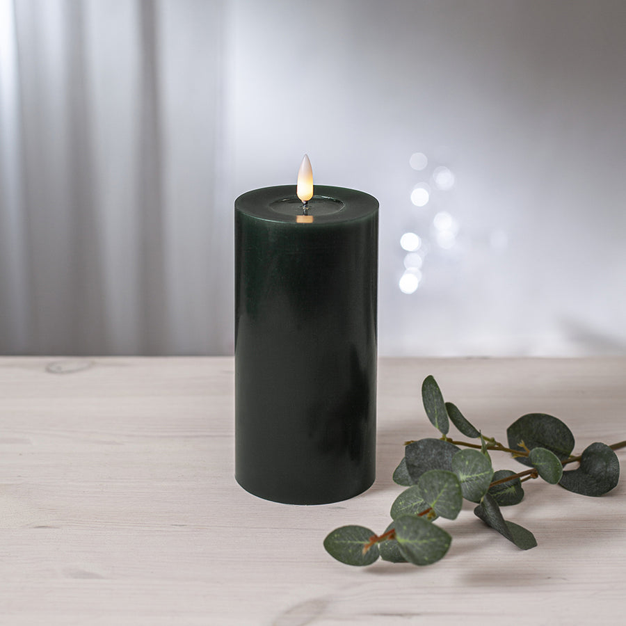 Zöld LED oszlopgyertya valódi viaszból, pislákoló lánggal, világos fa felületen, eukaliptuszlevéllel díszítve.