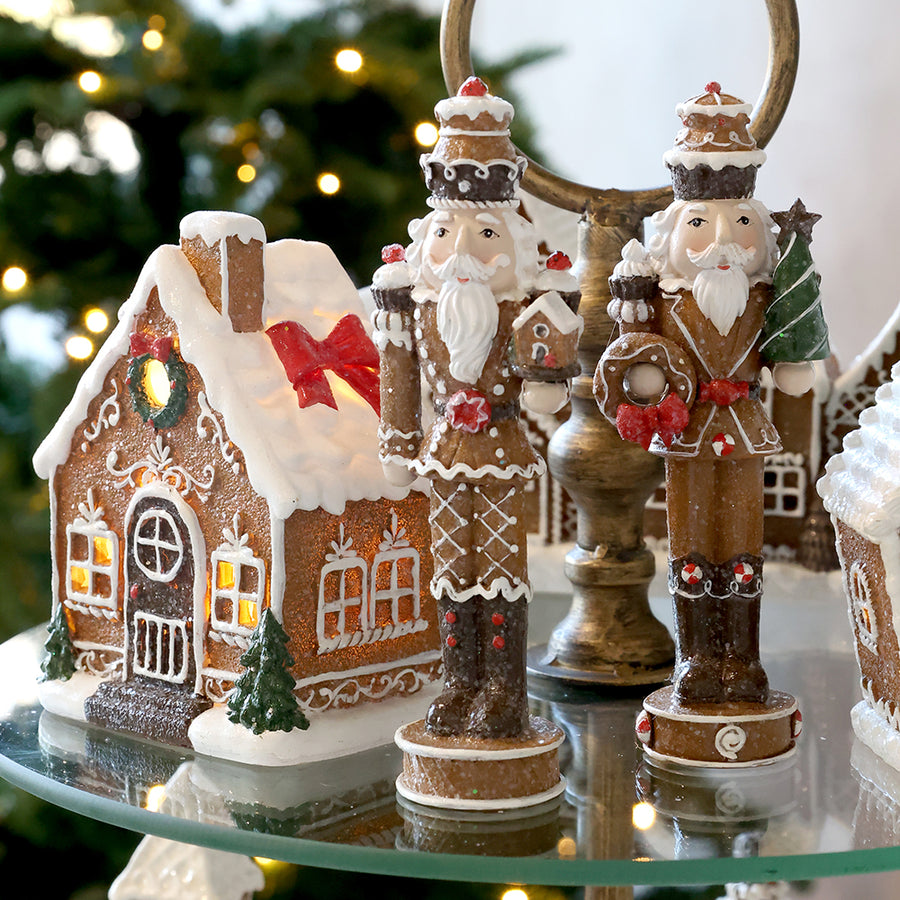 Gingerbread diótörő figurák üvegpolcon, világító mézeskalácsház mellett, háttérben karácsonyi fényekkel.