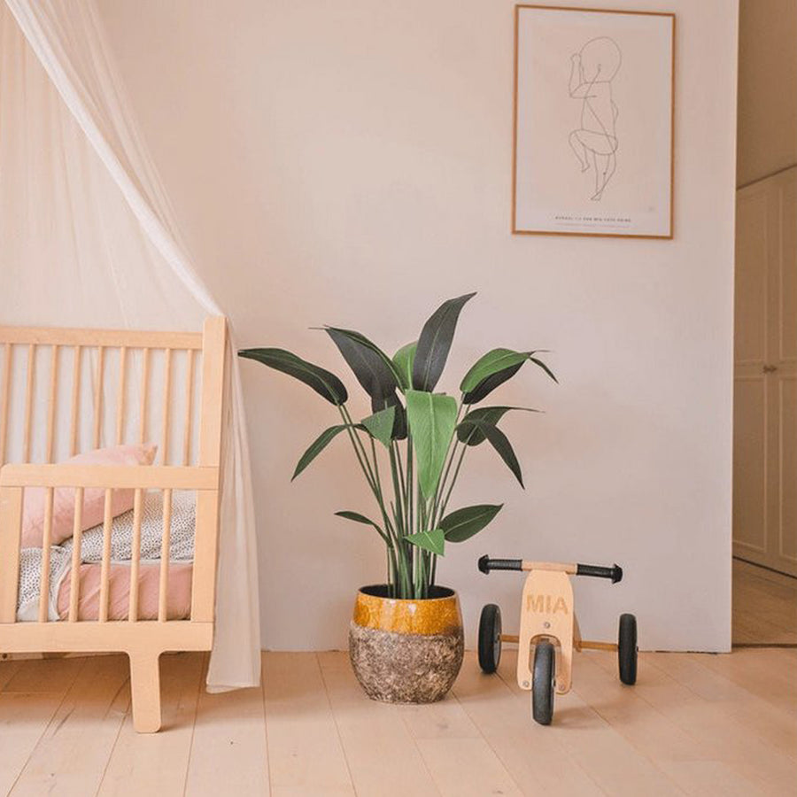Heliconia mű rákollóvirág dekoratív kaspóban, világos gyerekszobai enteriőrben kisággyal és fa játékbiciklivel