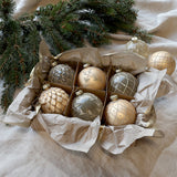 Pezsgőszínű üveg karácsonyfadísz szett rusztikus csomagolásban, fenyőágak mellett elhelyezve, Heritage kollekció.