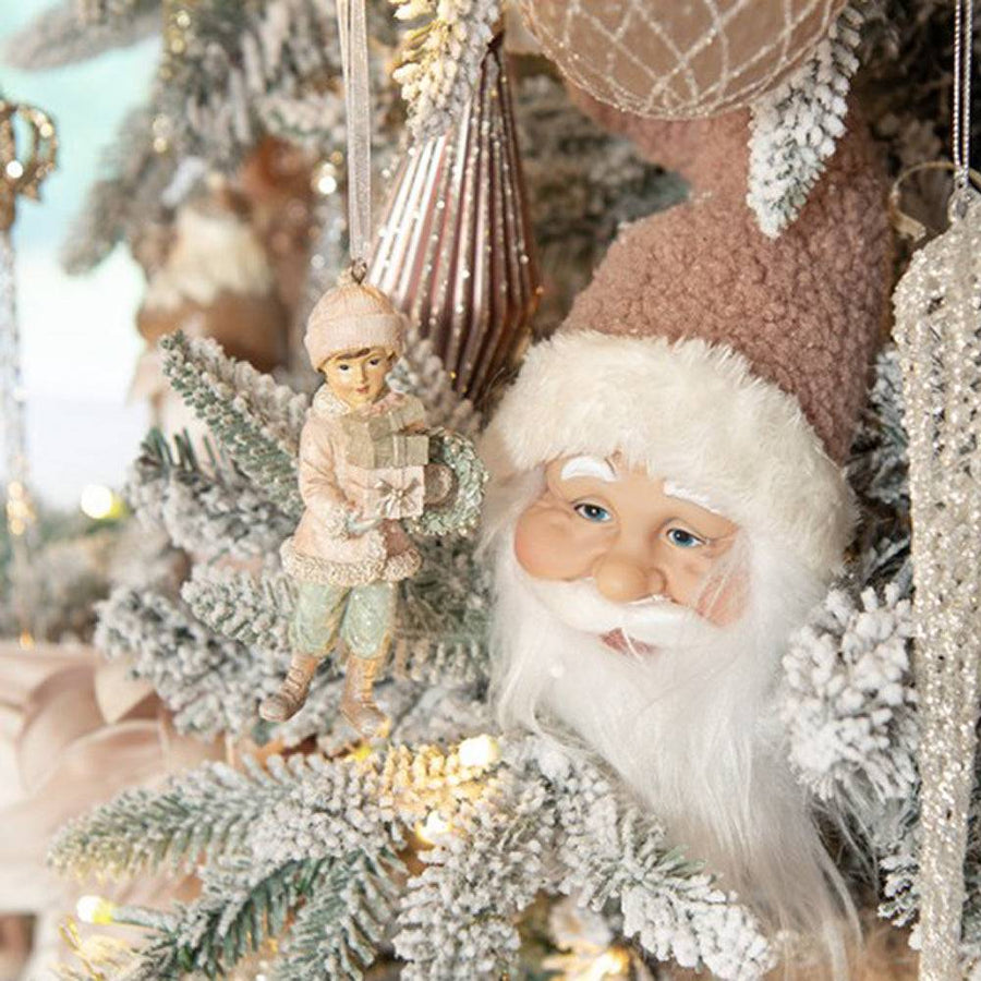 Heritage pink kisfiú karácsonyfadísz elhelyezve egy feldíszített, havas karácsonyfán, nosztalgikus dekorációban.