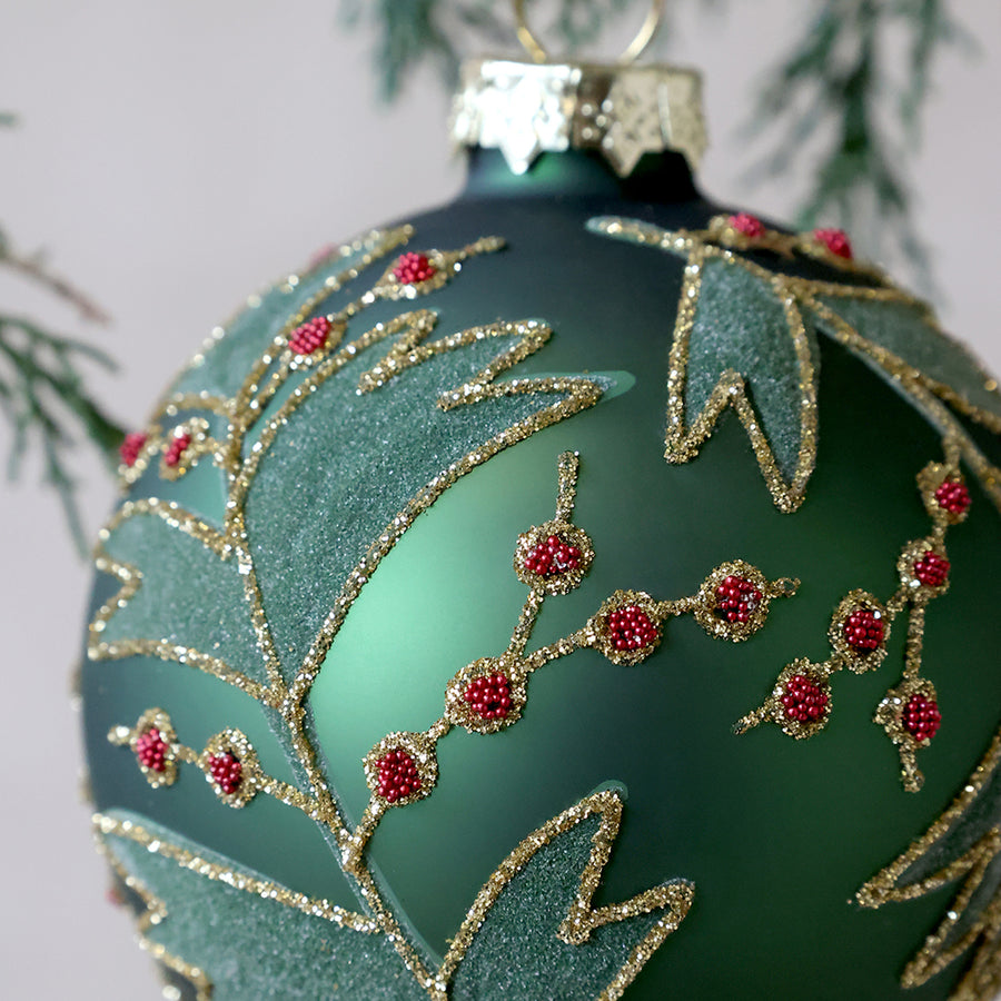 A Heritage zöld üveggömb csillámos levélmintájának és apró piros gyöngyökből álló bogyódíszítésének közeli részlete.