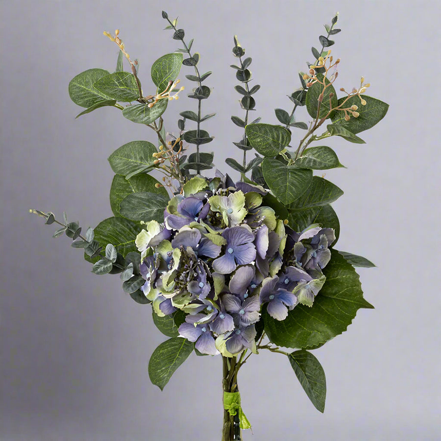 Kék és zöld árnyalatú mű hortenzia csokor szürke háttér előtt, elegáns zöld lombozattal.
