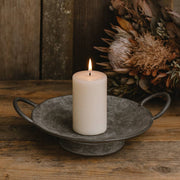 Égő gyertya a JUNKER szürke bádog tálban rusztikus fa felületen, szárított virágkompozícióval a háttérben.